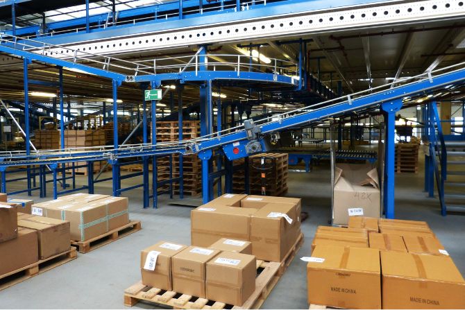 Cardboard boxes on pallets in a warehouse with conveyor systems, showcasing the importance of proper packaging and labeling for secure and efficient logistics.