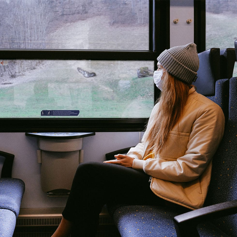 A woman wearing a surgical mask sitting in a train, looking outside the window.